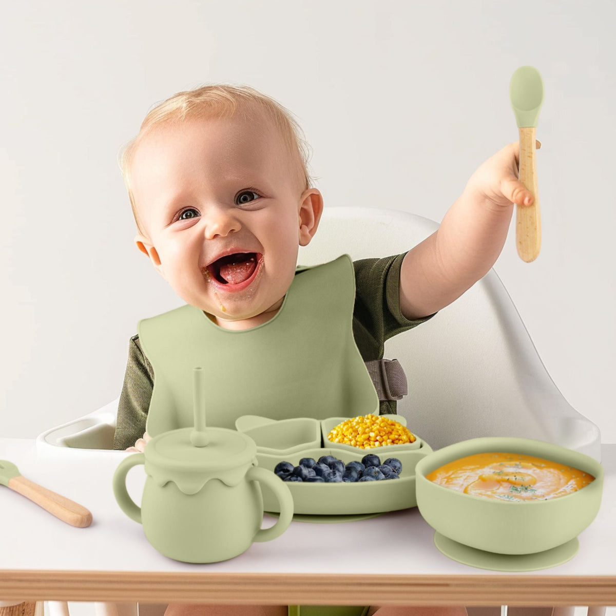 Baby in a high chair eating from a sage silicone suction bowl, divided plate and cup